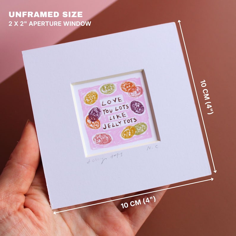 May include: A framed artwork with a pink background and colorful jelly tot illustrations. The text reads "LOVE YOU LOTS LIKE JELLY TOTS." The white frame measures 4 inches by 4 inches, with an aperture window of 2 x 2 inches.