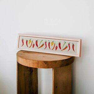 May include: A rectangular framed print featuring a row of colorful chili peppers. The peppers are in shades of red, orange, yellow, and green, set against a beige background. The frame is a light wood color, and the print is displayed on a wooden stool.