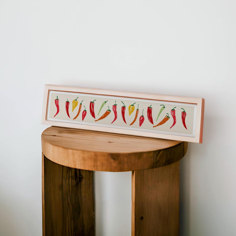 May include: A rectangular framed print featuring a row of colorful chili peppers. The peppers are in shades of red, orange, yellow, and green, set against a beige background. The frame is a light wood color, and the print is displayed on a wooden stool.
