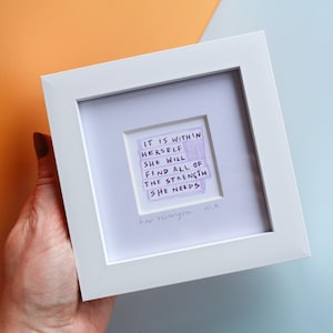 May include: A white framed art piece with a handwritten quote on a purple background. The quote reads: "IT IS WITHIN HERSELF SHE WILL FIND ALL OF THE STRENGTH SHE NEEDS." The art is held by a hand against a colorful background.
