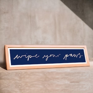May include: A rectangular wooden framed sign with the words "wipe your paws" in white script on a navy blue background. The sign is leaning against a neutral-coloured wall.