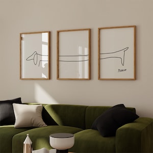 May include: Three framed black and white line drawings of a dachshund dog. The drawings are in a minimalist style and are hung on a beige wall above a green velvet sofa.