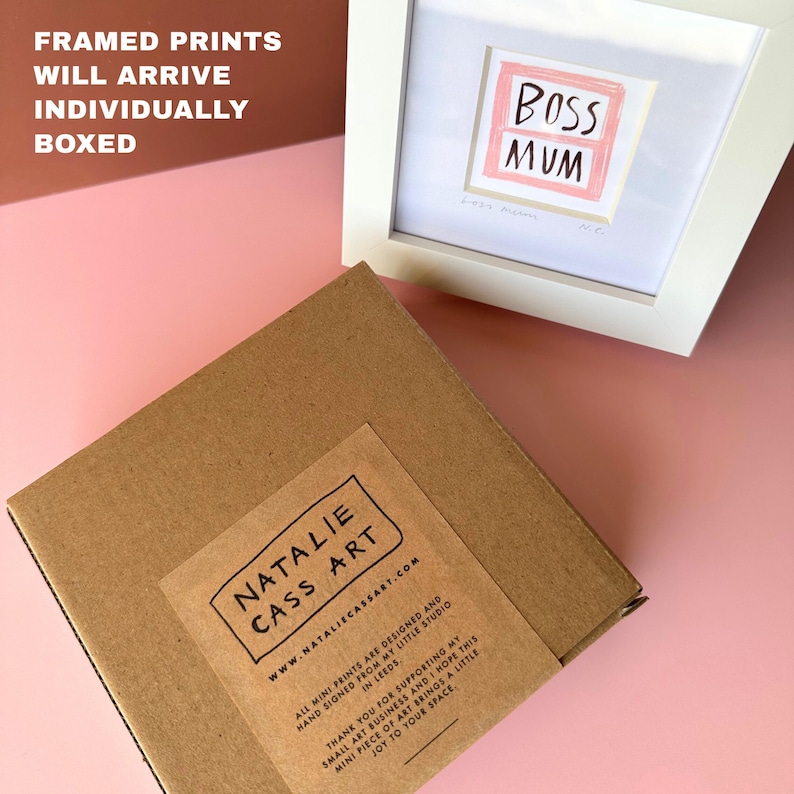 May include: A framed print with the words "BOSS MUM" in a pink and white design. The print is in a white frame, next to a brown cardboard box with a label that reads "NATALIE CASS ART". The text "FRAMED PRINTS WILL ARRIVE INDIVIDUALLY BOXED" is at the top.