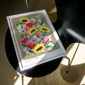 Op de afbeelding: Een ingelijste kunstdruk met een boeket zonnebloemen, rode klaprozen en andere kleurrijke bloemen. Het kunstwerk, gesigneerd met "Flower market", heeft een neutrale achtergrond en een witte lijst.