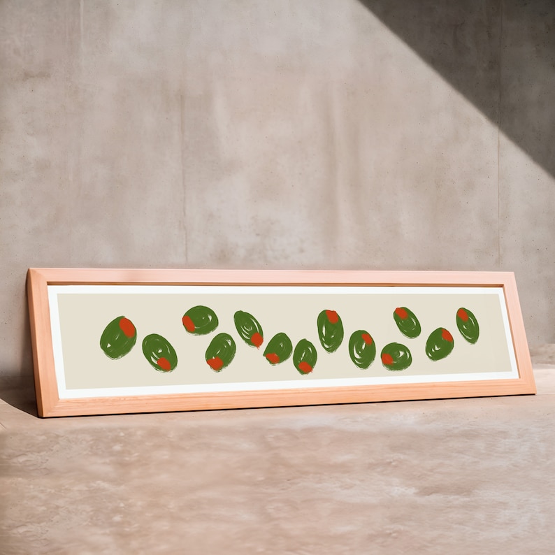 May include: A rectangular framed art print featuring a series of green olives with red centers, arranged on a cream-colored background. The frame is made of light-colored wood. The artwork is leaning against a neutral-toned wall.
