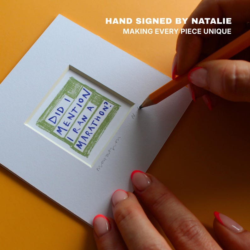 May include: A white art print with the text "DID I MENTION I RAN A MARATHON?" in blue and green. The print is being hand-signed with a pencil. The text "HAND SIGNED BY NATALIE MAKING EVERY PIECE UNIQUE" is also visible.