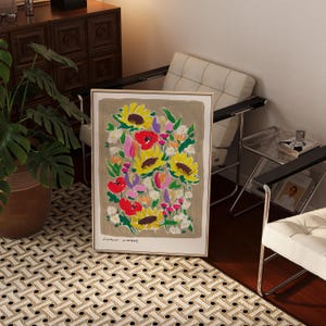 Op de afbeelding: Een ingelijste bloemenkunstprint met zonnebloemen, klaprozen en andere bloemen in geel, rood, roze en wit. De tekst "Flower Market" staat onderaan. Het kunstwerk wordt getoond op een patroonkleed.