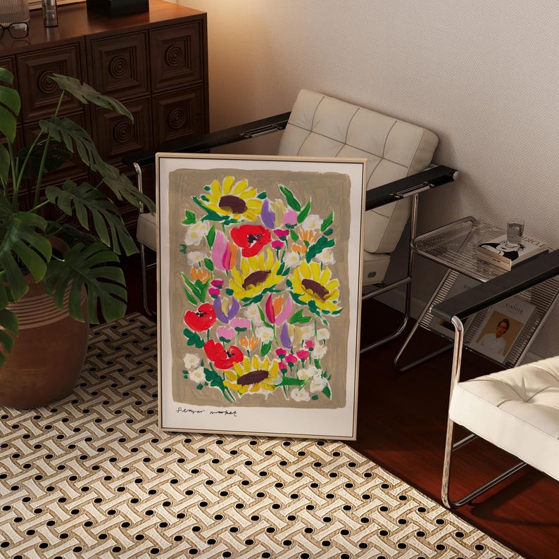 Op de afbeelding: Een ingelijste bloemenkunstprint met zonnebloemen, klaprozen en andere bloemen in geel, rood, roze en wit. De tekst "Flower Market" staat onderaan. Het kunstwerk wordt getoond op een patroonkleed.