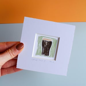 May include: A framed artwork featuring a hand-drawn illustration of a pint of stout. The artwork is set against a green background within a white frame. The text "spit the G N.C." is written at the bottom.