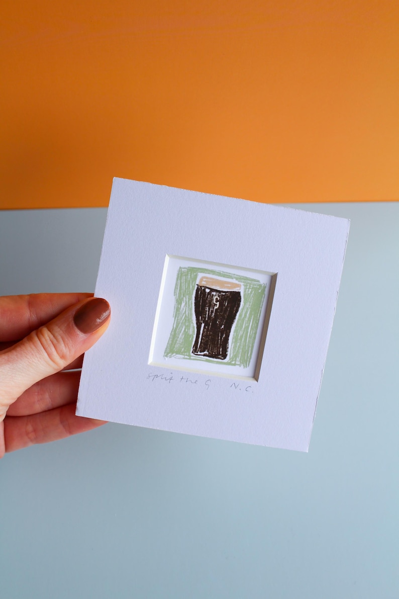 May include: A framed artwork featuring a hand-drawn illustration of a pint of stout. The artwork is set against a green background within a white frame. The text "spit the G N.C." is written at the bottom.