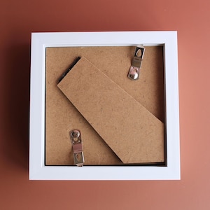 May include: A white square picture frame with a brown backing and metal clips. A brown cardboard stand is propped up against the back. The frame is sitting on a brown surface.