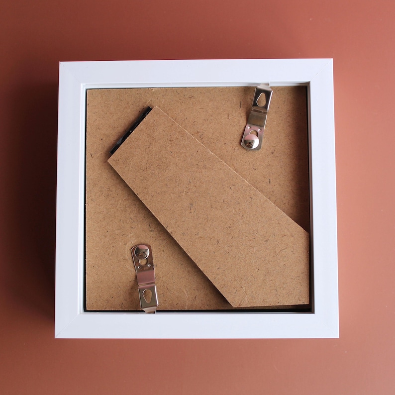 May include: A white square picture frame with a brown backing and metal clips. A brown cardboard stand is propped up against the back. The frame is sitting on a brown surface.