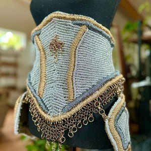 Könnte beinhalten: Ein handgefertigter silber- und goldfarbener Korsett, das auf einer schwarzen Schaufensterpuppe ausgestellt ist. Das Korsett weist aufwendige Webdetails, goldfarbene Verzierungen und einen Kettenfransenbesatz auf. Ein kleiner, dekorativer Anhänger hängt in der Mitte.