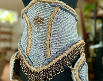 May include: A handcrafted silver and gold-toned corset displayed on a black mannequin. The corset features intricate woven details, gold trim, and a chainmail fringe. A small, decorative charm hangs from the center.
