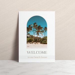 Beach house welcome sign featuring a minimalist design. A white card with a beige arched frame showcases a tropical scene: palm trees framing a light-blue beach house under a clear sky.  Text reads: 'WELCOME to our beach house' in a simple serif font. The overall aesthetic is calming and evokes a vacation vibe. Ideal for beach house decor or as a summer-themed printable.