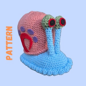 Puede incluir: Un juguete de caracol de crochet con una concha rosa, un cuerpo azul y ojos verdes. La concha tiene un diseño rojo y morado.