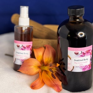May include: Two bottles of brown liquid with white labels that say "Snatched Body" and "Big mama's helper" on a white surface with an orange flower.