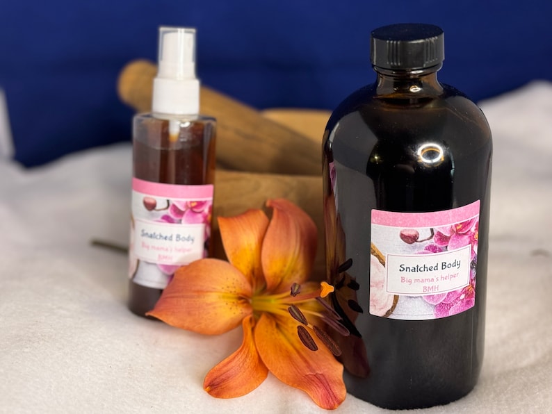 May include: Two bottles of brown liquid with white labels that say "Snatched Body" and "Big mama's helper" on a white surface with an orange flower.