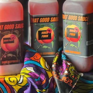 Può includere: Tre bottiglie di vetro trasparente con liquido rosso e tappi bianchi. Le bottiglie hanno etichette nere con testo bianco che recita "That Good Sauce" e "Hunch Punch". Le bottiglie sono appoggiate su un tessuto colorato a fantasia.