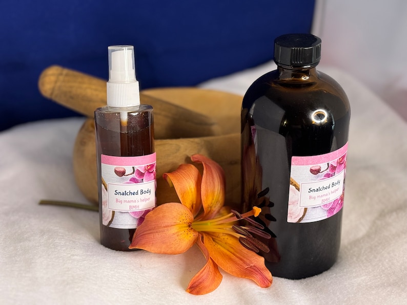 May include: Two brown glass bottles with labels that read "Snatched Body Big mama's helper BMH". The bottles are sitting on a white surface with a single orange lily flower.