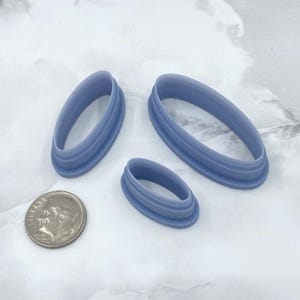 May include: Three light blue oval-shaped cookie cutters of varying sizes. A dime is next to the cutters for size comparison. The cutters have a ridged design on the sides. The cutters are made of plastic and are on a white surface.