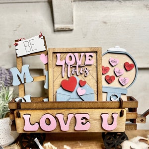 May include: A wooden wagon with a sign that says "Love Notes" and a gumball machine with hearts that say "Hugs", "XOXO", "Sweet", "Kisses", "Be Mine", and "I Love You". The wagon also has a sign that says "Love U" and a ladder with a sign that says "Be Mine".