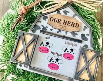 May include: A white and black barn-shaped Christmas ornament with the text "Our Herd 2022" on the front. The ornament has three cow silhouettes with the names "Jeff", "Danielle", and "Hudson" written below them.