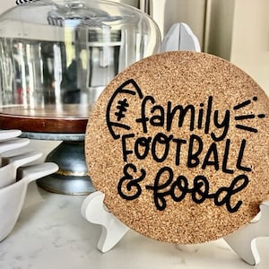 May include: A cork coaster with the text "#family=FOOTBALL & food" written in black cursive font. The coaster is on a white stand.