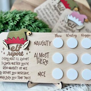 May include: Wooden elf report chart with a green elf hat and the text "elf report" and "EVERY NIGHT I'LL REPORT TO SANTA WHETHER YOU'VE BEEN NAUGHTY OR NICE. SO BEFORE YOU MISBEHAVE, MAKE SURE YOU THINK TWICE!"  The chart has three columns labeled "Theo", "Wesley", and "Reggie" with three white circles in each column labeled "NAUGHTY", "ALMOST THERE", and "NICE".