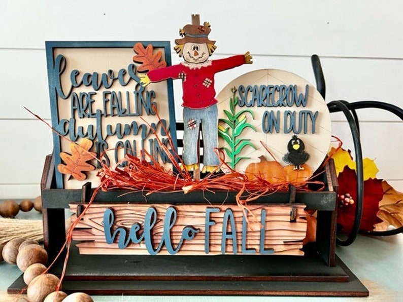 May include: A wooden fall-themed decorative piece with a scarecrow figure, pumpkins, and the text "Hello Fall." The piece includes a sign that reads "Leaves are falling, autumn is calling." and another sign that reads "Scarecrow on Duty."