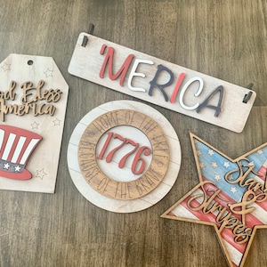 May include: Four wooden signs with a patriotic theme. The signs feature the phrases "God Bless America", "'MERICA", "Home of the Free Because of the Brave 1776", and "Stars & Stripes". The signs are painted in red, white, and blue.
