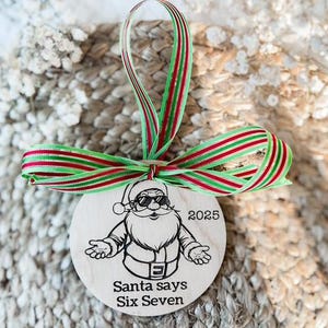 May include: A round wooden Christmas ornament featuring a cartoon Santa Claus wearing sunglasses. The ornament has the text "2025" and "Santa says Six Seven". It is adorned with a red and green striped ribbon.