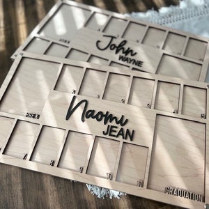 May include: A wooden photo frame with 15 slots for pictures. The frame is light brown and has the words "Naomi Jean" and "GRADUATION" engraved on it.