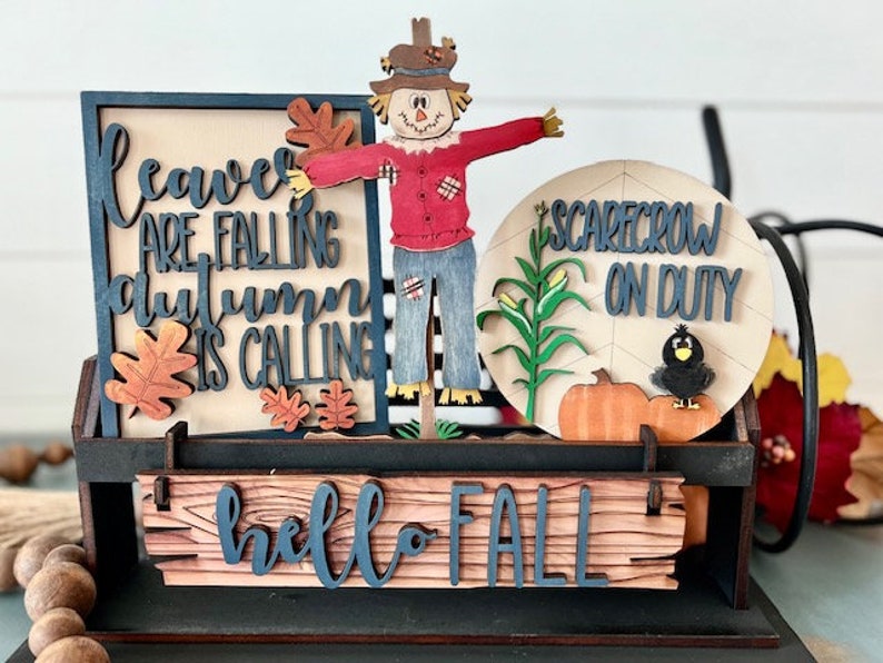 May include: A wooden fall-themed decoration featuring a scarecrow, pumpkins, and the phrases "Leaves are falling, autumn is calling" and "Hello Fall." The scarecrow wears a red shirt and blue jeans. A black crow sits on a pumpkin.