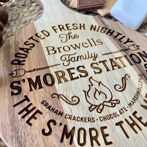May include: A round wooden cutting board with a handle, engraved with the text "Roasted Fresh Nightly The Browells Family S'mores Station The S'more The Merrier Graham Crackers Chocolate Marshmallows".
