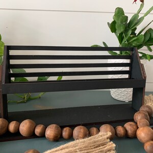 May include: A black wooden shelf with horizontal slats, designed to hold items. The shelf is set against a backdrop of greenery and a white pot. A string of wooden beads and a jute tassel are in the foreground.