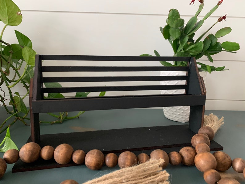 May include: A black wooden shelf with horizontal slats, designed to hold items. The shelf is set against a backdrop of greenery and a white pot. A string of wooden beads and a jute tassel are in the foreground.