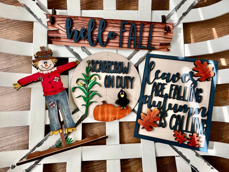 May include: Wooden fall-themed decorations, including a sign that says "hello FALL", a scarecrow, and a sign that says "Leaves are falling autumn is calling". The decorations are in shades of red, orange, brown, and blue.