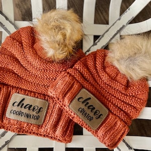 May include: Two orange knit beanies with faux fur pom-poms. One beanie has a leather patch that reads "chaos COORDINATOR" and the other reads "chaos CREATOR". The beanies are sitting on a white, distressed, wooden surface.