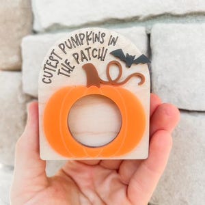 May include: Wooden Halloween decoration featuring an orange pumpkin with a cutout center and a brown stem. The text "Cutest Pumpkins in the Patch!" is above the pumpkin, with a small bat silhouette.