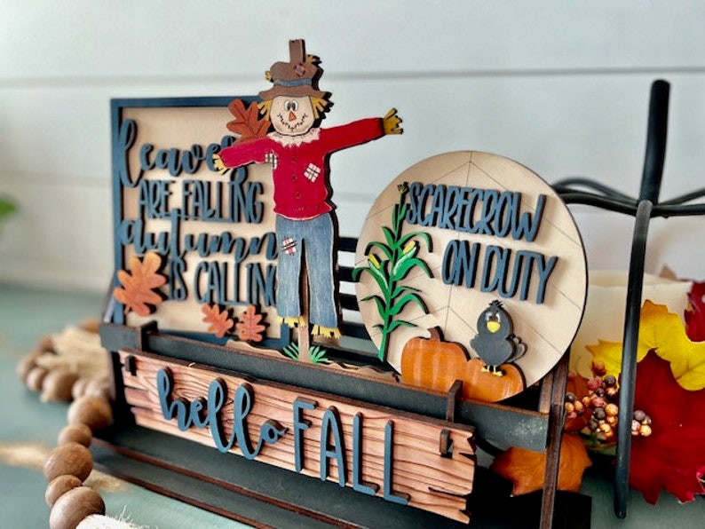 May include: A wooden fall-themed decoration featuring a scarecrow figure with outstretched arms. The sign includes the phrases "leaves are falling, autumn is calling" and "hello fall". A circular sign reads "scarecrow on duty".