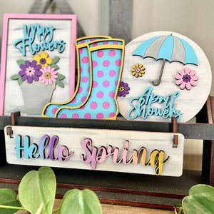 May include: A wooden spring-themed tiered shelf with three signs. The top sign says "May Flowers" with a pink background and a bouquet of purple and pink flowers. The middle sign says "April Showers" with a blue umbrella and a white background. The bottom sign says "Hello Spring" with a rainbow gradient.