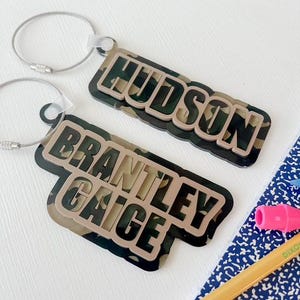 May include: Two personalized name tags with a camouflage design. The names "HUDSON" and "BRANTLEY GAGE" are displayed in a bold font. Each tag has a silver-colored metal loop attached for easy hanging.