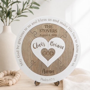 May include: A circular wooden puzzle plaque featuring a white border with the text May God be gracious to us and bless us and make His face to shine upon us. The center includes the names The Stovers, August 8, 2026, Chris, Briana, Aaron, and God.