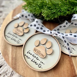 May include: Three wooden ornaments with paw prints and the text "forever in our hearts" and the names "Nico", "Willow", and "[unclear name]" on a wooden surface. The ornaments are tied with white ribbon with black paw print designs.