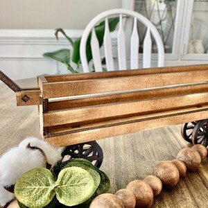 May include: A wooden toy wagon with a pull handle and spoked wheels. The wagon is made of light brown wood and has a rectangular shape. Decorative elements include a cotton ball and wooden beads.