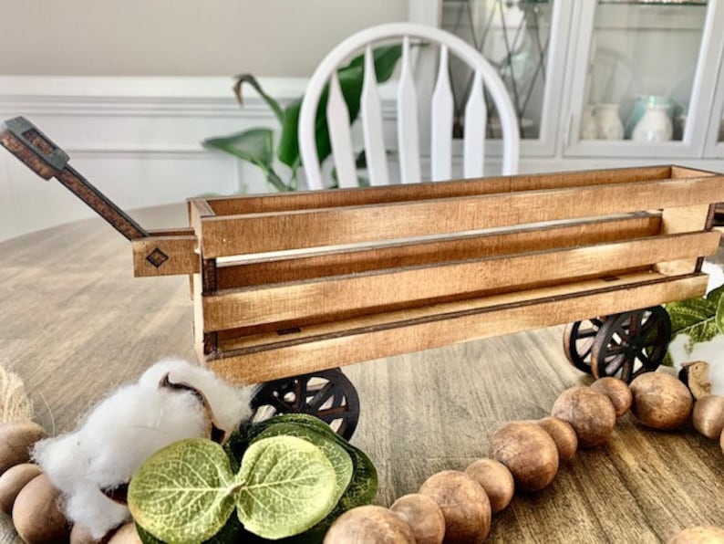 May include: A wooden toy wagon with a pull handle and spoked wheels. The wagon is made of light brown wood and has a rectangular shape. Decorative elements include a cotton ball and wooden beads.