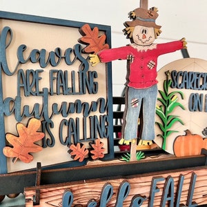 May include: Wooden fall-themed decoration featuring a scarecrow, pumpkins, and the text "Leaves are falling, autumn is calling." The design includes a cart with a sign that says "Hello Fall."