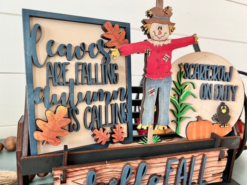 May include: Wooden fall-themed decoration featuring a scarecrow, pumpkins, and the text "Leaves are falling, autumn is calling." The design includes a cart with a sign that says "Hello Fall."