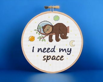 May include: A white embroidery hoop with a cross-stitch design of a sloth wearing an astronaut helmet and sleeping on a branch. The design includes stars, planets, and a crescent moon. The text "I need my space" is stitched below the sloth.
